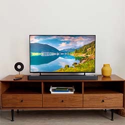 A black vinyl record player with an open transparent lid sits on a wooden cabinet. A record is spinning, and a small vase with green leaves is nearby, creating a retro ambiance.