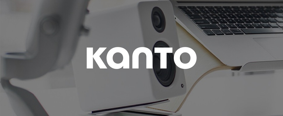 Modern workspace with a laptop on a wooden stand, flanked by speakers. A hand writes with a stylus, conveying a minimalist and tech-savvy vibe. "Kanto" written in bottom left