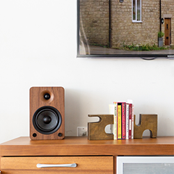 Wooden speaker and books with geometric bookends on a light wood cabinet, under a wall-mounted TV displaying a brick building. 