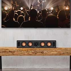 A sleek black speaker with copper accents sits on a wooden shelf. Above, a flat-screen TV displays a lively concert with a crowd and bright stage lights.