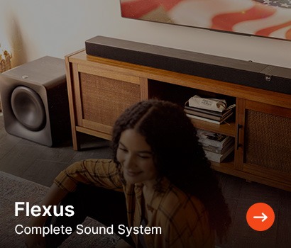 A woman with curly hair sits on the floor, smiling near a modern wooden entertainment unit featuring a sleek soundbar and a subwoofer. "Flexus Complete Sound System" text overlay.