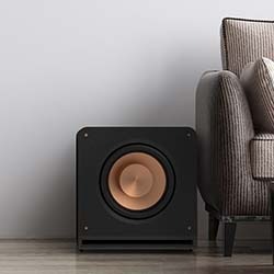 A sleek black subwoofer with a copper-coloured speaker cone is placed on a wooden floor next to a modern, textured, beige armchair against a light grey wall.