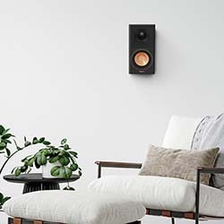 A minimalist living room with a white chair and ottoman, a grey pillow, green plant on a low table, and a black speaker mounted on a white wall.