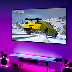 A video game scene on a wall-mounted screen shows a yellow car drifting on a snowy road. The room is lit with vibrant purple LED lights, creating an immersive atmosphere.