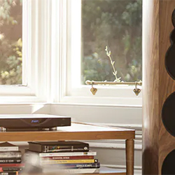 Cozy room corner with books stacked on a wooden table, a sleek speaker nearby. Sunlight filters through a window, creating a warm and inviting atmosphere.
