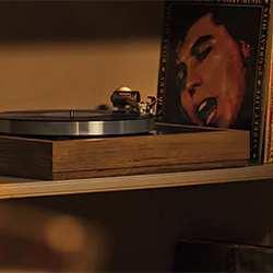 A vintage wooden turntable sits on a shelf, spinning a vinyl record. An album cover with a close-up of a singer's face is propped up beside it, lit warmly.