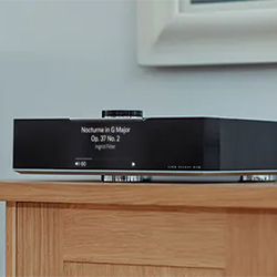A sleek black digital music player on a wooden shelf displaying "Nocturne in G Major Op. 37 No. 2" on its screen, set against a light blue wall.