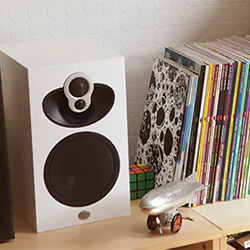 A white speaker and a silver toy airplane sit on a shelf next to a colourful stack of vinyl records and a Rubik's Cube, creating a retro-modern vibe.