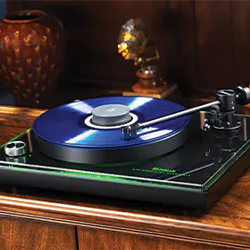 A blue vinyl record spins on a sleek modern turntable with a glowing edge, set atop a polished wooden surface. The room has a cosy, vintage feel.