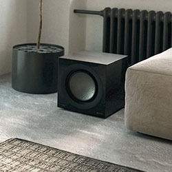 A modern, minimalistic living room corner featuring a black cube-shaped speaker near a grey sofa and a black radiator, with neutral tones and a calm ambiance.