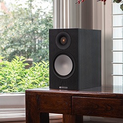 A black speaker with a large grey woofer and small tweeter sits on a wooden table by a window, with greenery visible outside, creating a serene setting.