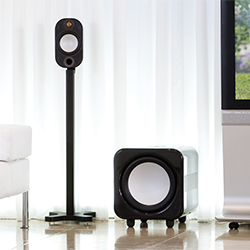 A modern home audio setup features a tall black speaker on a stand and a cube-shaped subwoofer on the floor, against white curtains in a sleek living room.