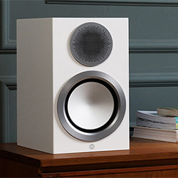 A modern white speaker with a silver-rimmed woofer and mesh tweeter, placed on a wooden table against a gray paneled wall, beside stacked books.