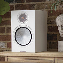 A white bookshelf speaker with a metallic driver and circular design sits on a wooden shelf against a brick wall. 