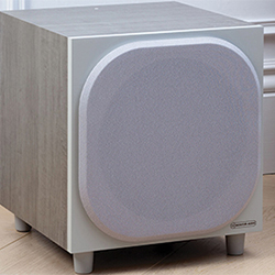 A modern gray subwoofer with a cube shape and a rounded front grille sits on a light wooden floor, exuding a sleek and minimalist design.