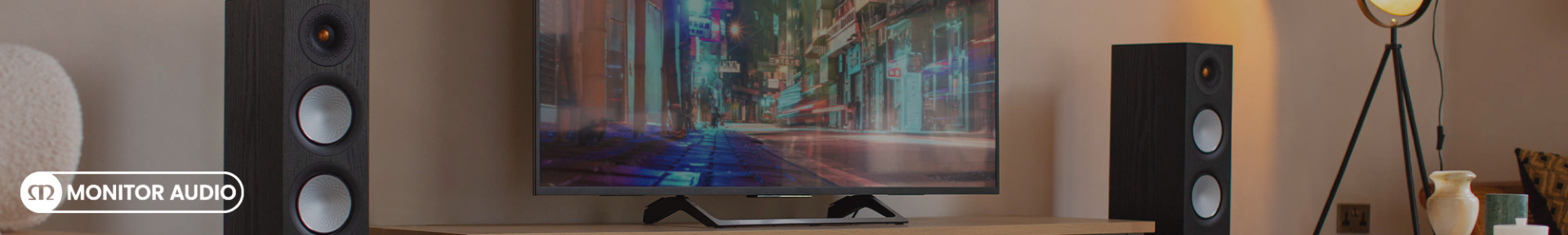 A sleek home entertainment setup features a large TV displaying a blurred city scene. Flanked by two tall black Monitor Audio speakers, the room is warmly lit, creating a cozy ambiance.