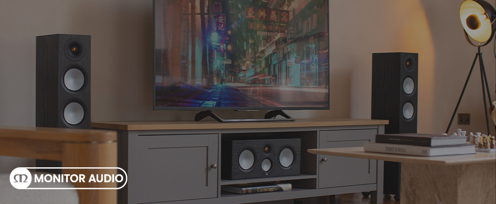 A sleek home entertainment setup features a large TV displaying a blurred city scene. Flanked by two tall black Monitor Audio speakers, the room is warmly lit, creating a cozy ambiance.