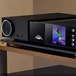 Black audio system with a large circular knob on the left and a lit digital display showing colourful graphics on the right. Placed on a wooden shelf.