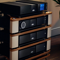 A sleek audio equipment rack with four components, each featuring a minimal design. The top unit has a display screen. The room is dimly lit, adding elegance.