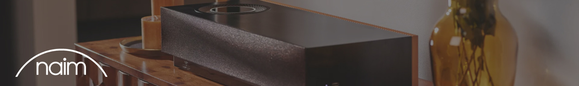 A sleek black speaker on a wooden table, next to a lit candle and amber glass vase. The word "naim" is in white text on a curved line in the corner.