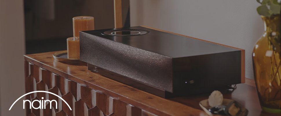 A sleek black speaker on a wooden table, next to a lit candle and amber glass vase. The word "naim" is in white text on a curved line in the corner.
