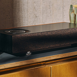 A sleek, rectangular black speaker sits on a wooden cabinet. A small cactus is beside it, and the concrete wall background gives a modern vibe.
