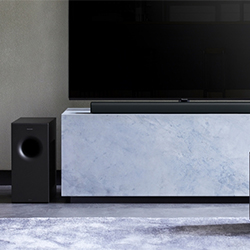 A sleek black soundbar sits in front of a large flat-screen TV on a marble stand, with a black subwoofer to the left, creating a modern home theater setup.