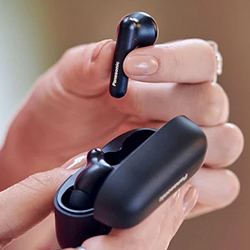 A hand holds a black earbud above its charging case, which is also held in the other hand. The setting is softly lit, conveying a sense of modern technology.