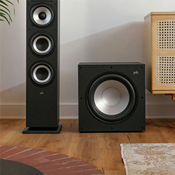 Floor-standing black speaker and a subwoofer placed on a wooden floor next to a cabinet. 