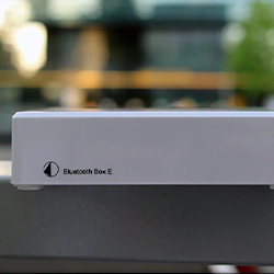 A white electronic device labelled "Bluetooth Box E" sits on a table. The background is blurred, with hints of greenery, suggesting an outdoor setting.