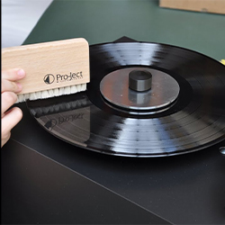 A person is using a wooden brush labelled "Pro-Ject" to clean a black vinyl record on a turntable. The setting is calm and focused on maintenance.