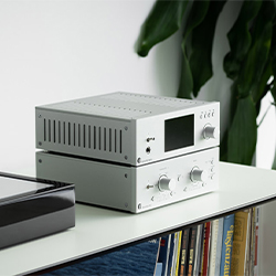 Silver audio amplifiers stacked on a white shelf with books. A large, leafy plant is blurred in the background, creating a modern and sleek ambiance.