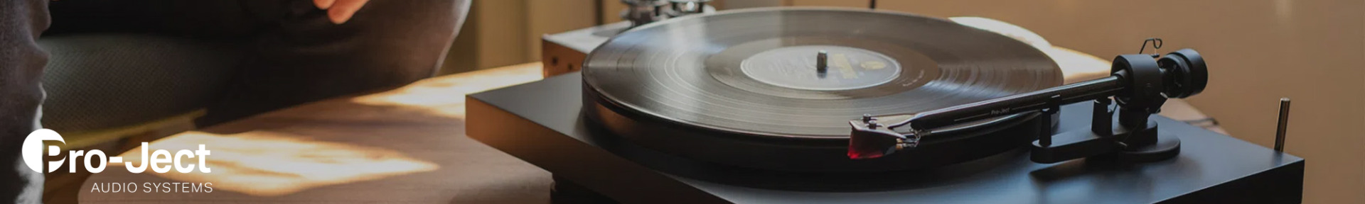 A vintage turntable with a spinning vinyl record sits in soft, warm lighting, conveying a nostalgic, relaxed atmosphere. The Pro-Ject Audio Systems logo is visible.