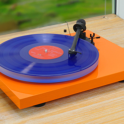 A vibrant orange turntable with a blue vinyl record spins on a wooden surface. Sunlight and greenery are visible through a nearby window, creating a calm atmosphere.