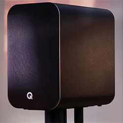A sleek, black speaker with a rounded rectangular design and a "Q" logo on the front, placed on a stand. The background is softly blurred.