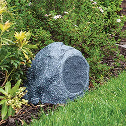 A realistic grey fake rock speaker is placed among lush green bushes and grass in a garden setting, blending naturally with the outdoor environment.