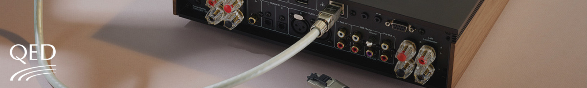 Back of an audio system showing various connectors and cables connected to ports. A coiled wire is prominent. The QED logo is visible on the left.