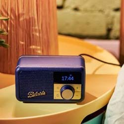 Retro-style blue radio with digital clock display reading 17:44, placed on a light-coloured table. Warm lighting and a cosy ambiance are present.