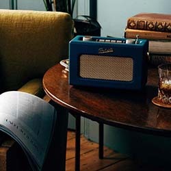A cosy room with a vintage blue radio on a wooden table, accompanied by stacked books and a glass of whiskey. A newspaper lies on a nearby green chair.
