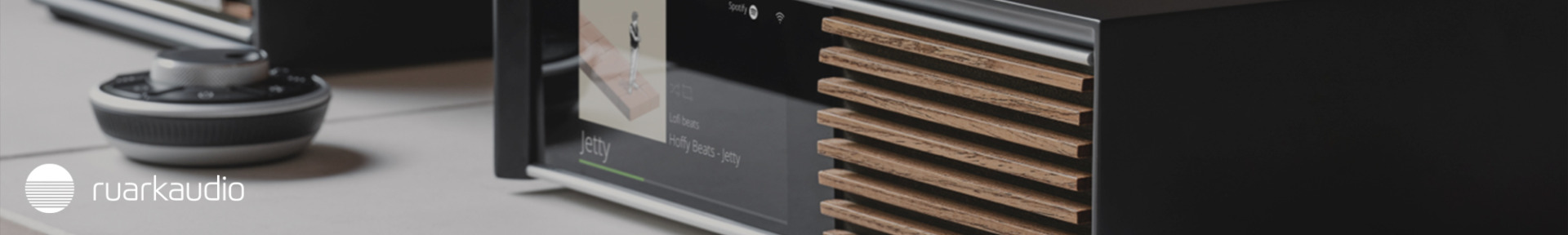Close-up of a modern audio system with wooden slats and a digital display showing a track titled "Jetty." A sleek turntable is visible in the background. Logo says "Ruark Audio." 