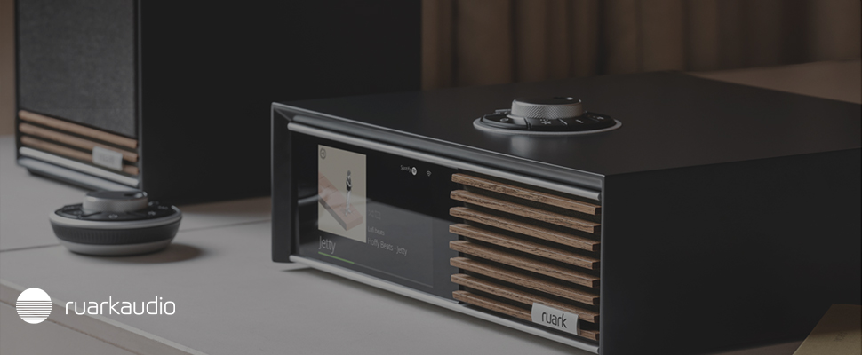 Close-up of a modern audio system with wooden slats and a digital display showing a track titled "Jetty." A sleek turntable is visible in the background. Logo says "Ruark Audio." 