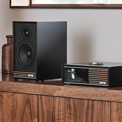 A black speaker and audio system sit on a wooden shelf. The modern, sleek design contrasts with the rustic shelf, creating a cosy, vintage ambiance.