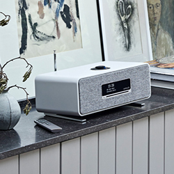 A sleek, modern radio with a digital display sits on a dark countertop. Behind it are framed abstract artworks, creating a minimalist and artistic vibe.