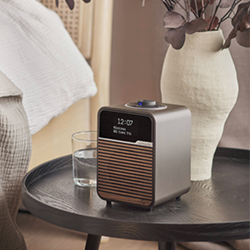 A modern, compact clock radio with a wooden front sits on a round table, next to a vase and a glass of water. 