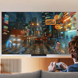 A person sits on a couch playing a racing video game on a large TV, displaying a vibrant, futuristic cityscape with neon lights and a dynamic atmosphere.