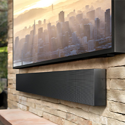 A sleek black soundbar is mounted on a stone wall below a TV displaying a hazy city skyline at sunset. The scene conveys a modern and peaceful ambiance.