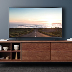 A sleek, modern TV displaying a scenic road leading to the sunrise, sits atop a wooden cabinet with open shelves holding decorative items. Calm, inviting scene.