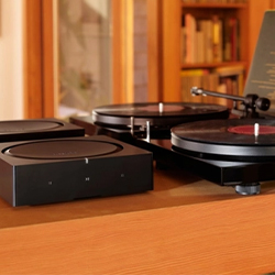 Turntable with a black vinyl record on a wooden surface, surrounded by speakers. Shelves of books in the background create a cosy, retro atmosphere.