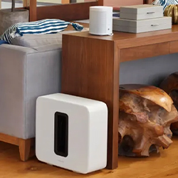 A white rectangular speaker sits on the floor beside a wooden table and a grey couch. Two beige boxes and a striped pillow are on the table, creating a cosy setting.