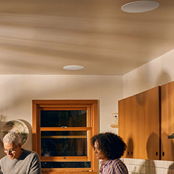 A couple is cooking in a warm-lit kitchen, smiling and engaged. Wooden cabinets and a window are visible, creating a cosy, inviting atmosphere.